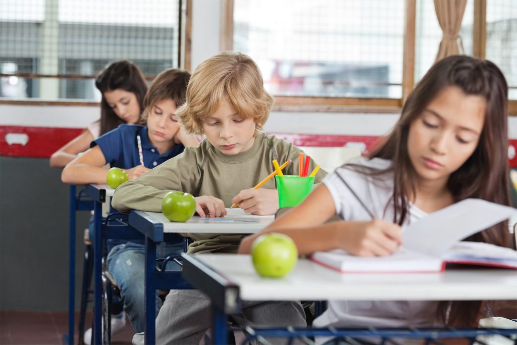 Kinder sitzen im Klassenzimmer und sind in Ihre Hefte vertieft. Auf jedem Tisch liegt ein Apfel.